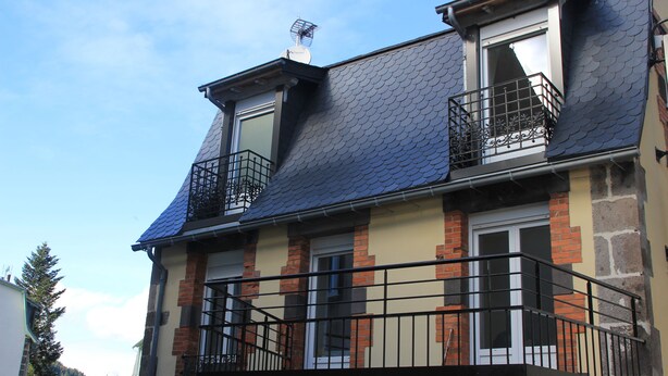 Um edifício de dois andares com telhado escuro, fachada de tijolo e reboco, e uma varanda com guarda-corpo de metal.