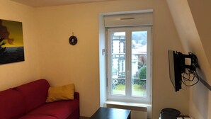 Quarto pequeno e bem iluminado com um sofá vermelho, uma mesa preta e uma janela com vista para edifícios e uma varanda.