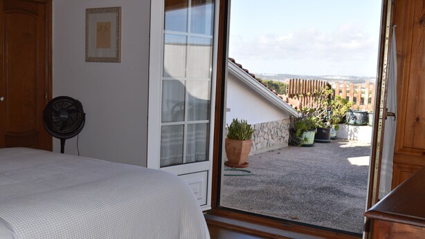 A bedroom with a bed, a fan, and a view of the outside balcony with potted plants. - Roof Top apartment, quiet, 2 large terraces, close to the best beaches (Lourinha)
