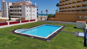 Une piscine entourée d’une pelouse bien entretenue, avec vue sur une plage et un immeuble à plusieurs étages en arrière-plan.