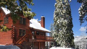 Una cabaña de troncos con balcón, rodeada de árboles cubiertos de nieve y un cielo azul despejado.