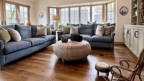 A spacious living room with a large sectional sofa, a round woven ottoman, and a wooden floor. There are large windows providing natural light and a ceiling fan. - 4-bedroom vacation home just 12 minutes from LEGOLAND® California (Oceanside)