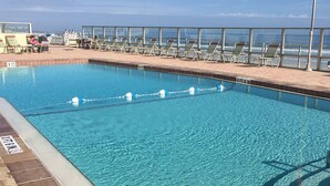 Una piscina azul cristalina con indicador de profundidad de 1.7 metros, rodeada de sillas de descanso y una barrera de vidrio con vista al océano.