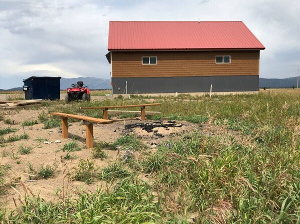 A building with a red roof and a black lower section, a fire pit, and a bench in a grassy field.
