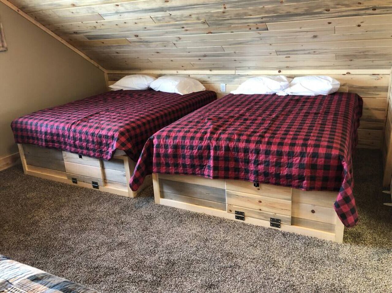 Een kamer met twee bedden, elk met een rood-zwart geruit sprei en witte kussens. De kamer heeft een schuin plafond met houten balken en een vloerbedekking.