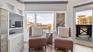 Een moderne kamer met twee bruine fauteuils, een kleine tafel en een televisie op een wit kastje. Grote ramen bieden uitzicht op een gebouw en een hek.