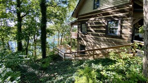 Eine rustikale Blockhütte mit Holzterrasse und kleiner Veranda, umgeben von dichtem Wald und einem See.