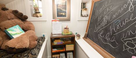Una habitación acogedora con un gran osito de peluche, una estantería llena de libros y juegos de mesa, una pizarra con un mensaje de bienvenida y una ventana que da a unas vistas arboladas.
