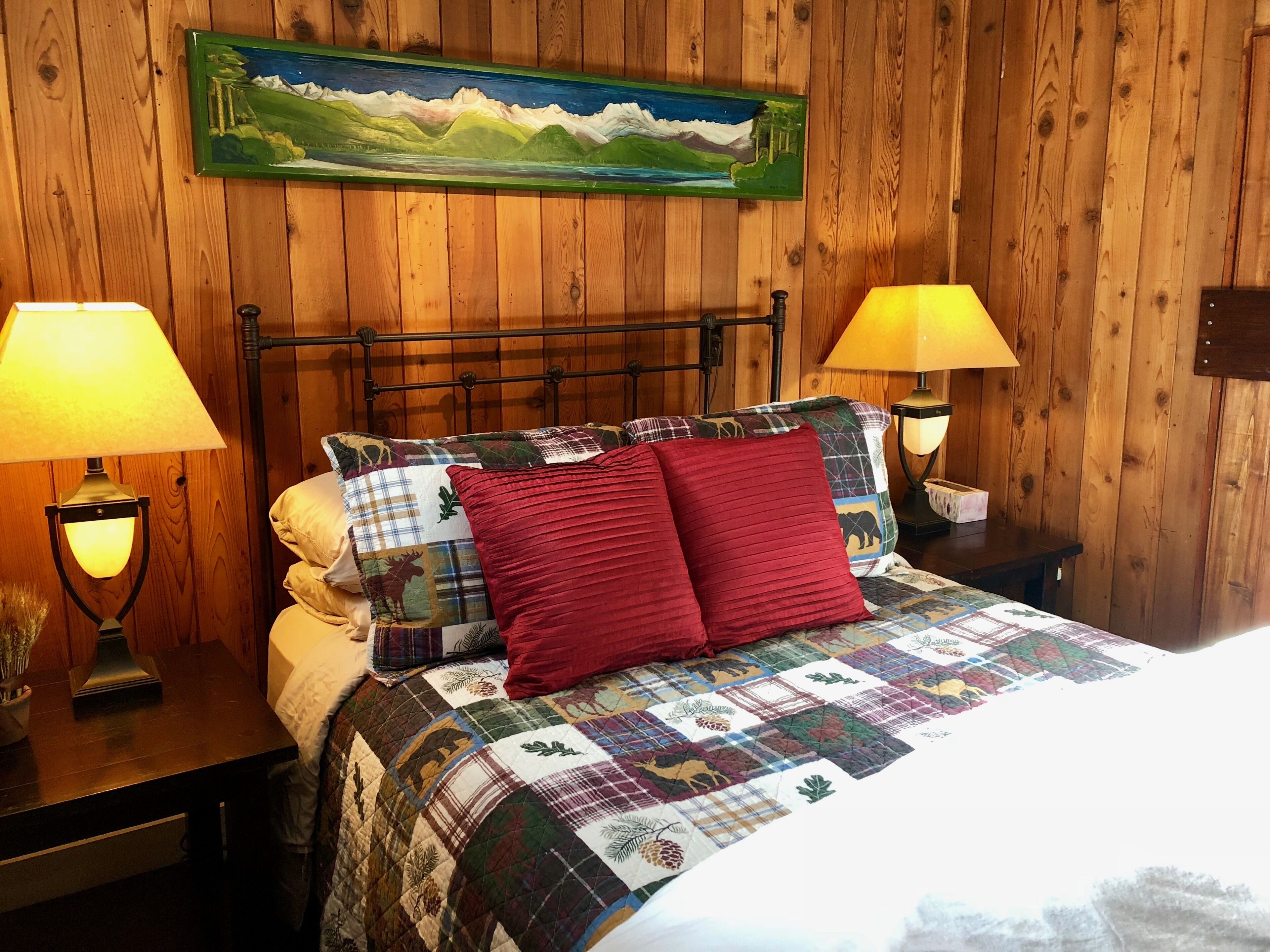 A wooden-paneled room with a bed, two bedside lamps, a quilted bedspread, and a painting on the wall.