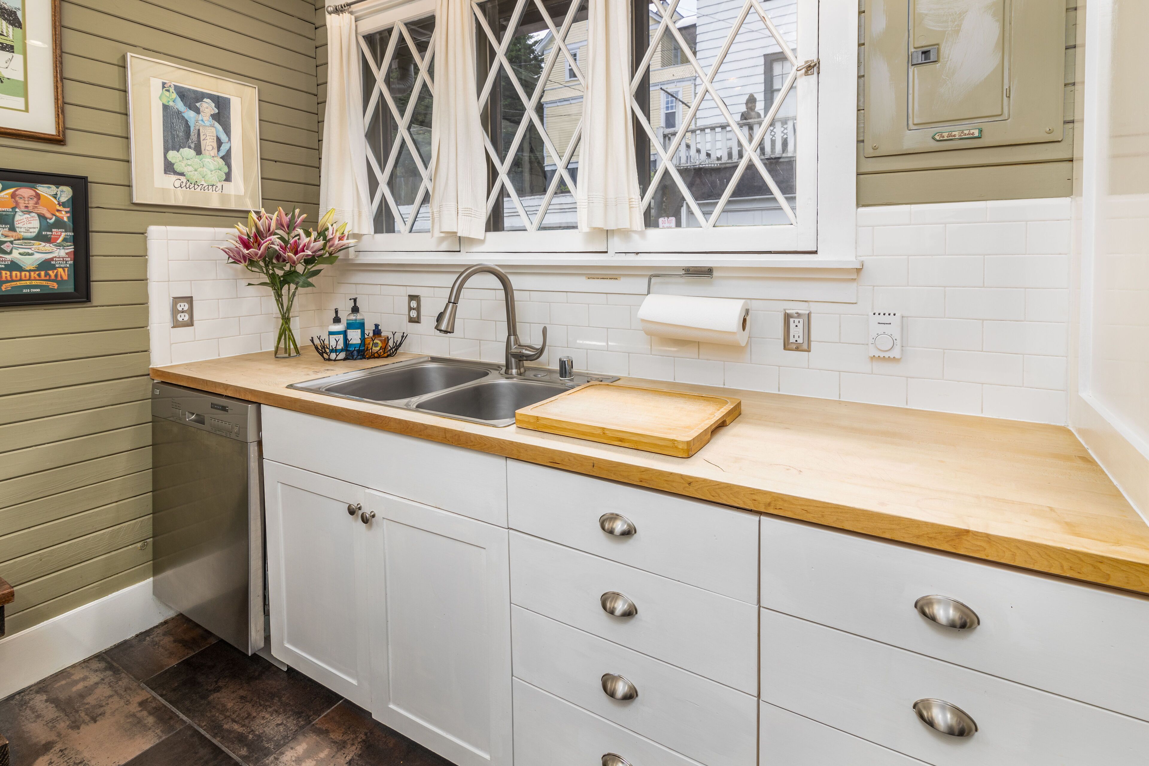 A kitchen with a sink, wooden countertop, white cabinets, and a window with white curtains.