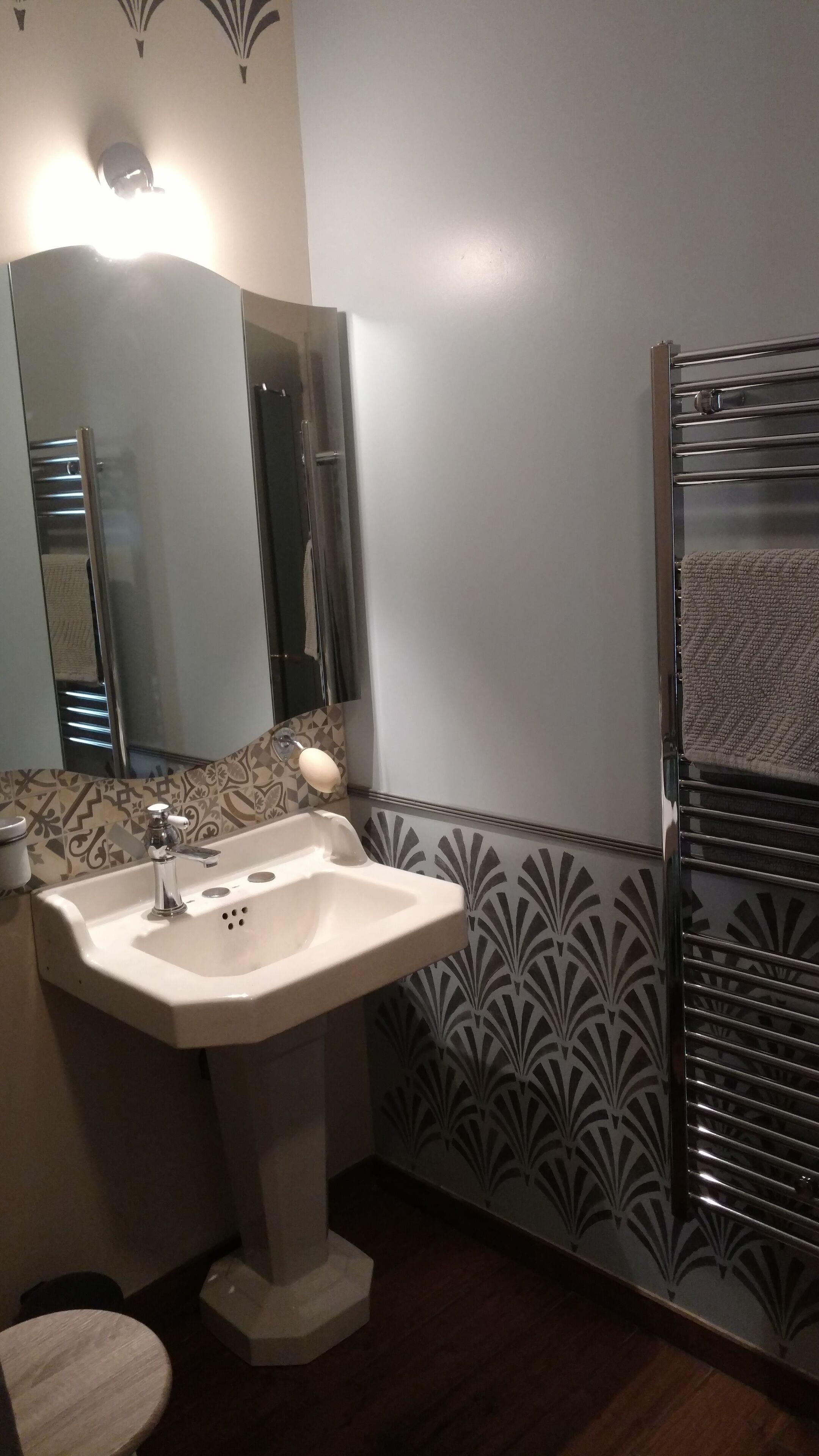 Een badkamer met een wastafel op een voetstuk, een spiegel, een handdoekhouder en betegelde wanden met een patroon.
