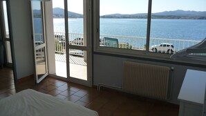 Ein Zimmer mit großem Fenster mit Blick auf ein Gewässer und Berge. Es gibt ein Bett, eine Heizung und einen Balkon mit Geländer.