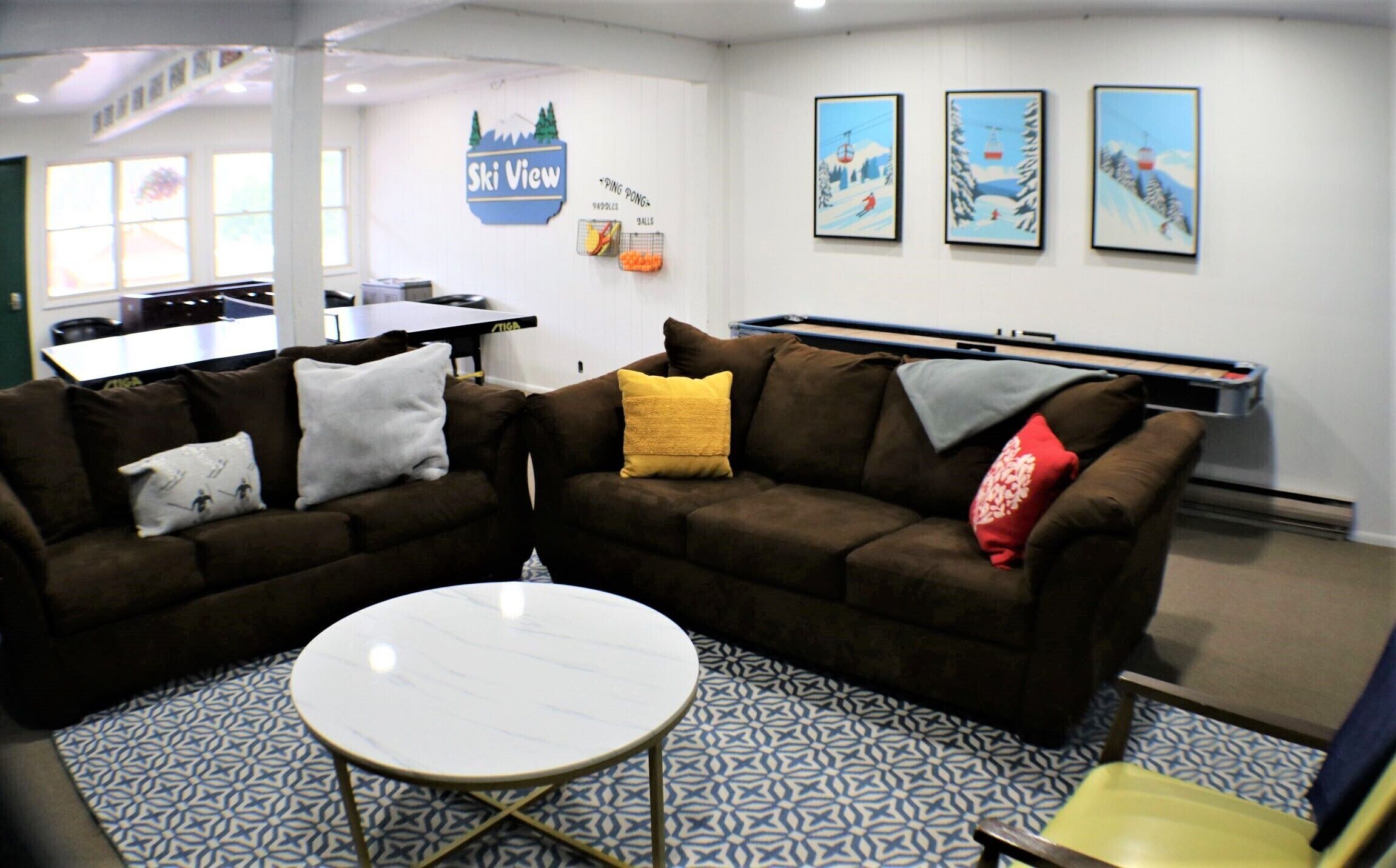 A room with two brown sofas, a white coffee table, patterned floor tiles, a ping pong table, and wall art depicting a ski scene.