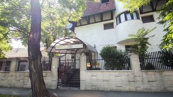 Entrada de hotel con un camino de acceso empedrado, un gran árbol y un edificio de fachada blanca y detalles oscuros.