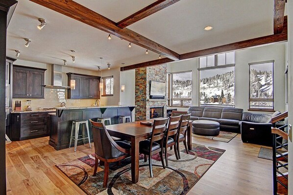 A spacious living area with a wooden floor, a dining table, and a sofa. There is a kitchen with dark cabinets and a large island. The room has large windows with a view of a snowy landscape.