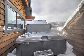 A hot tub outside a wooden cabin with a snowy mountain view.