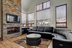 A modern living room with a stone fireplace, leather sofa, and large windows offering a snowy mountain view. - On Main St, rooftop hottub, porch w/ covered grill, loft,walk to gondola (Breckenridge)