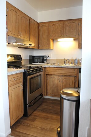 A kitchen with wooden cabinets, a stainless steel stove, a microwave, and a trash can. - Bright End Unit Apartment (Anchorage)