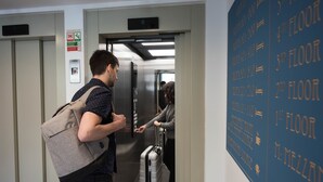 Un pasillo con un ascensor, una pared con indicaciones de pisos y un cartel de alarma de incendios.