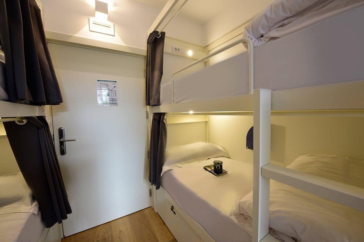 Habitación con literas, una puerta, cortinas y una pequeña mesa con una cámara.