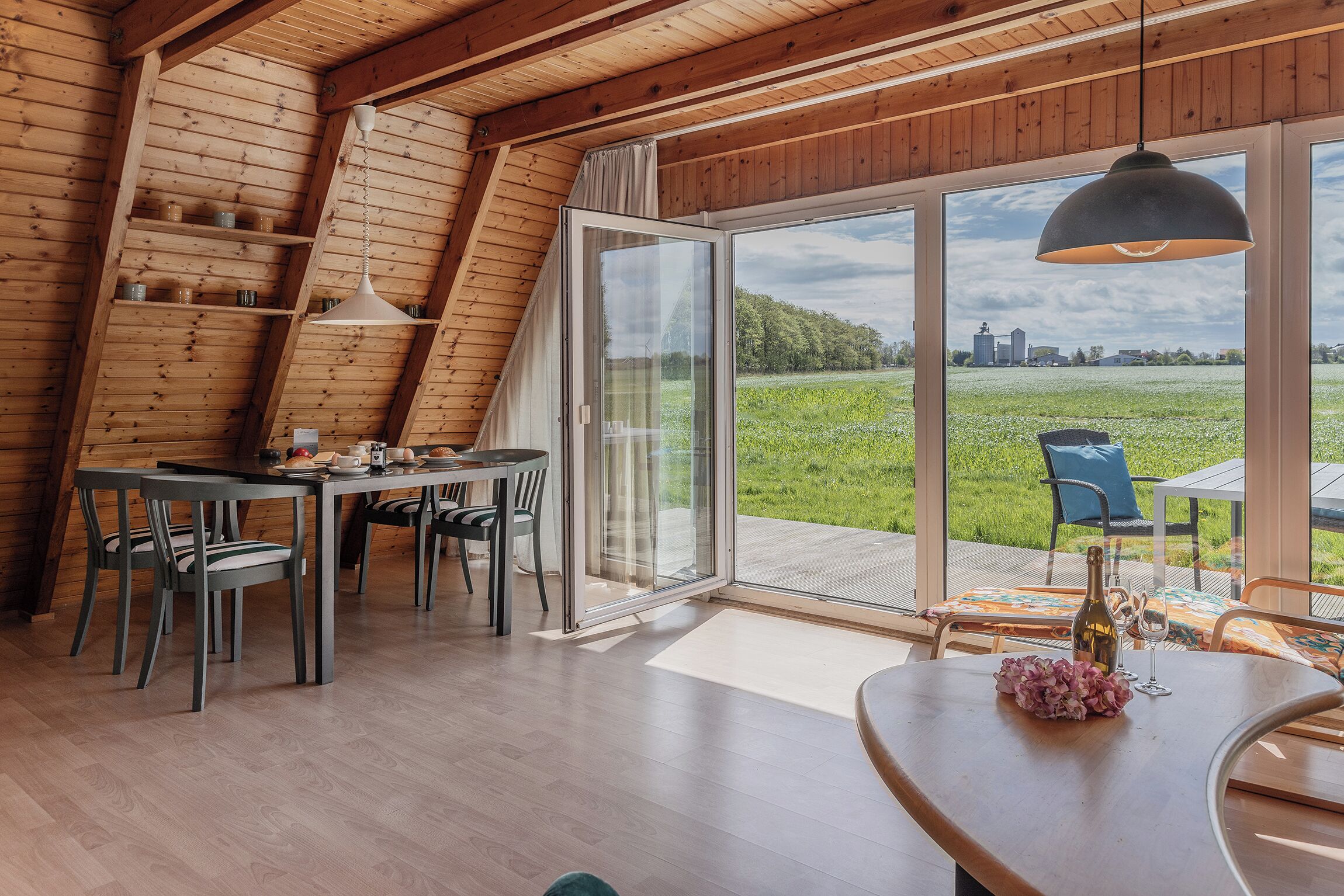 Un espace repas comprenant une table et des chaises, une grande fenêtre donnant sur un champ verdoyant et des poutres en bois au plafond.