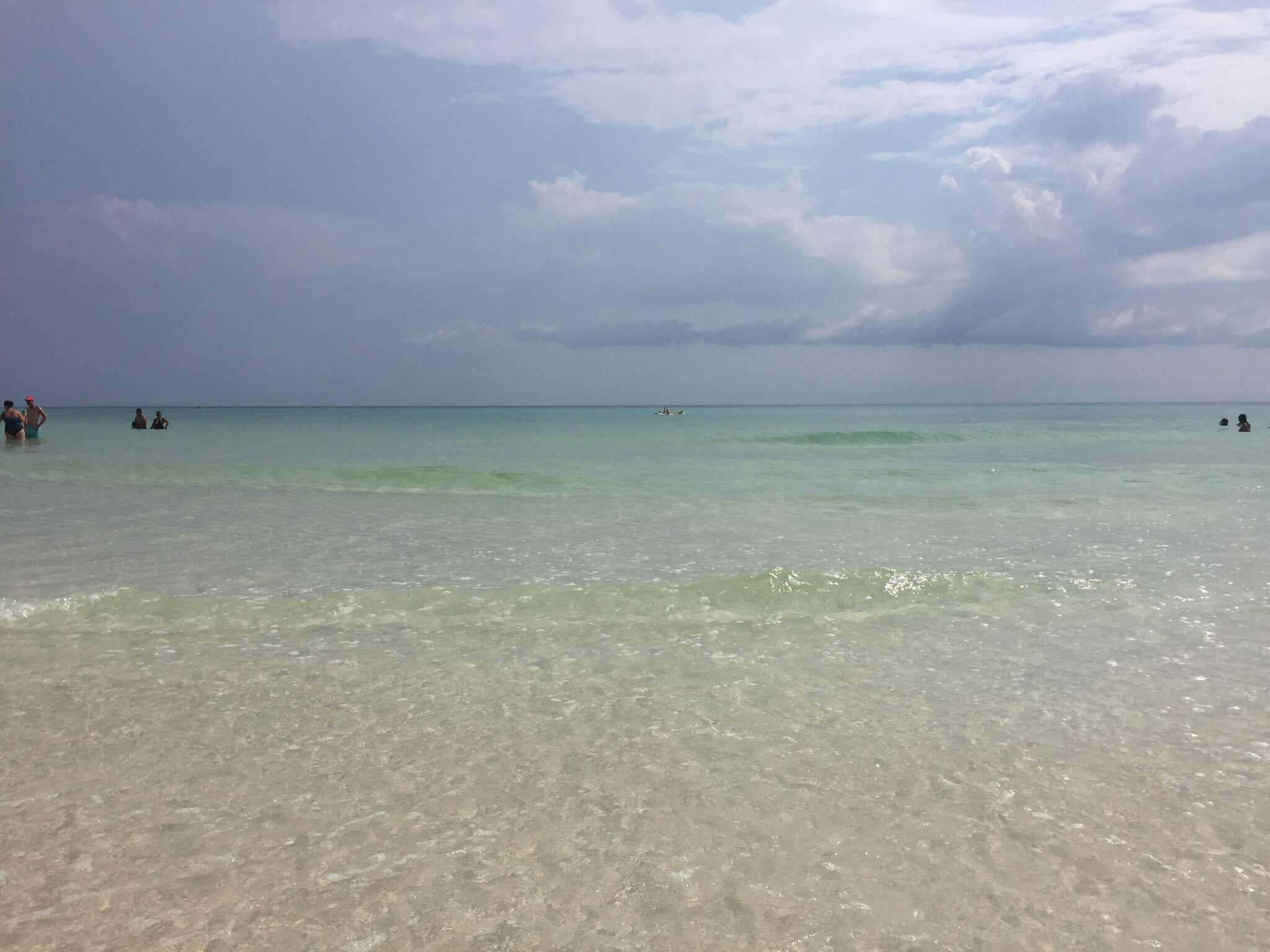 A beach with clear shallow water, a few people in the distance, and a cloudy sky.