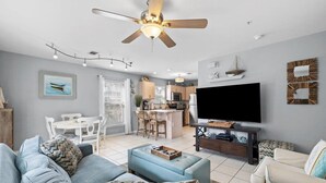 A modern living room with a sofa, ottoman, and a TV on a stand. There is a ceiling fan, a dining area with a table and chairs, and a kitchen in the background. - Modern coastal 1st floor 30A unit w/ walkability to restaurants & beach! (Santa Rosa Beach)