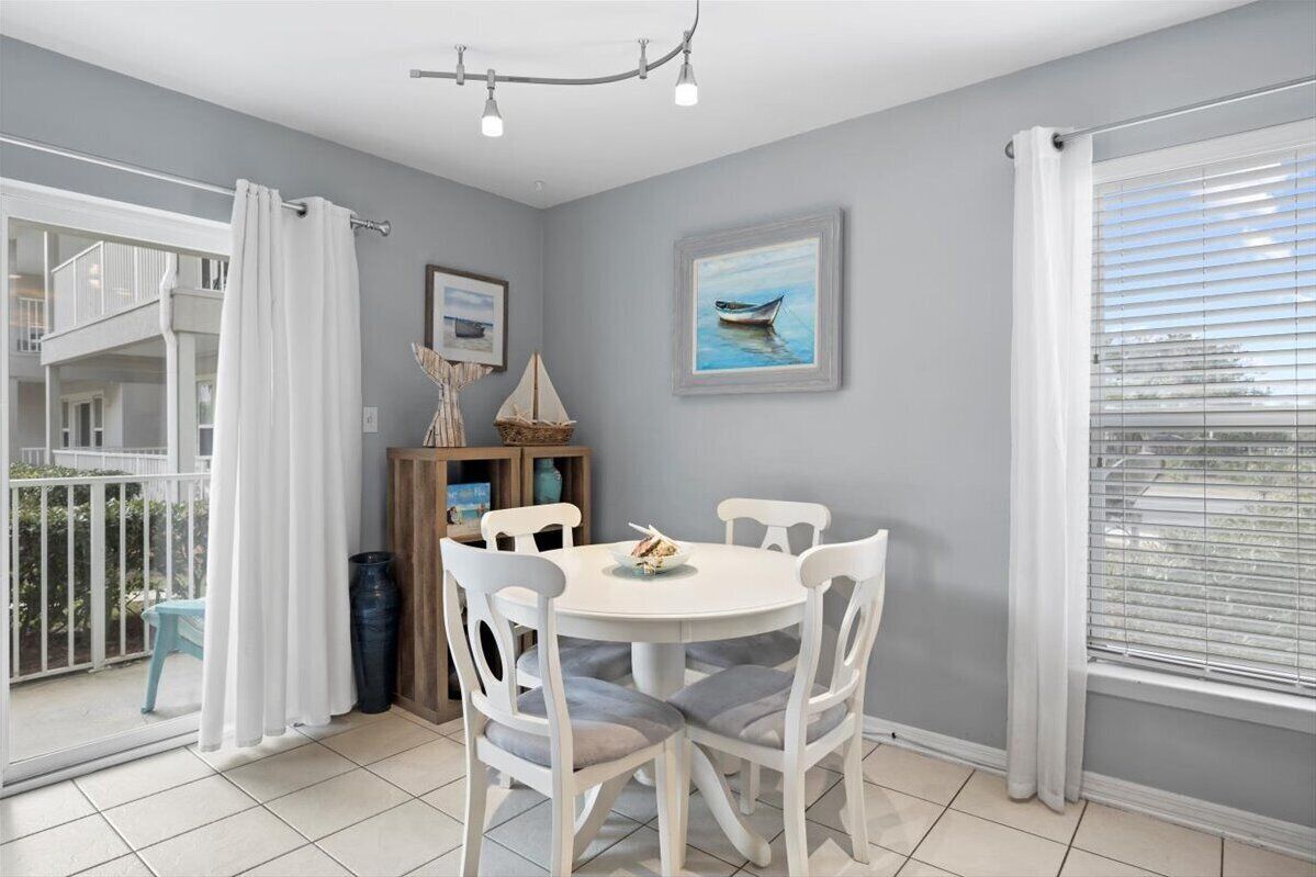 A dining area with a round table and white chairs, a framed picture on the wall, and a window with blinds.