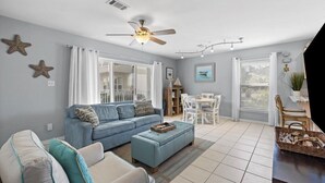 A living room with a blue sofa, a ceiling fan, and a dining area with a table and chairs. - Modern coastal 1st floor 30A unit w/ walkability to restaurants & beach! (Santa Rosa Beach)