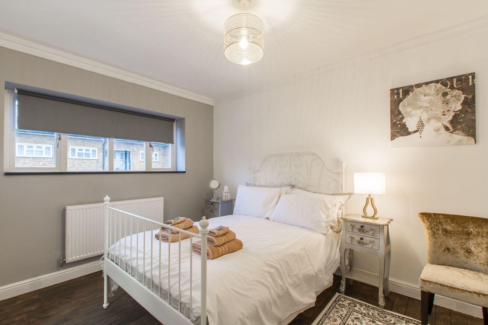 Ett sovrum med en säng, ett element, ett fönster, en lampa, en stol och en tavla på väggen.