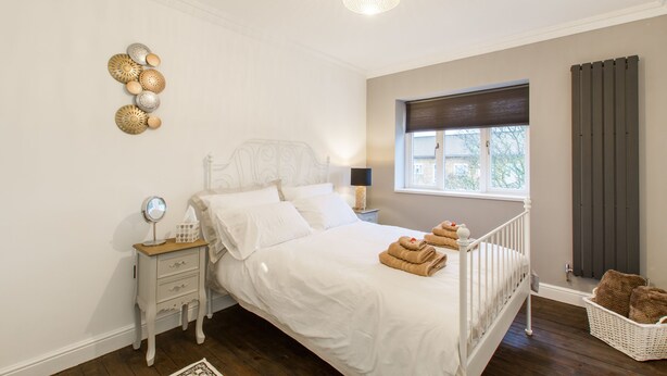 A bedroom with a white bed, a nightstand with a mirror, a lamp, and a window with blinds. - Beautiful Luxury London Flat - 3 minutes from Piccadilly line - sleeps 12 (London)