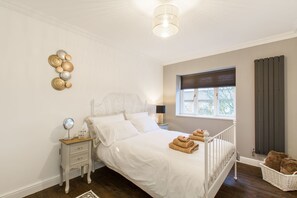 Ein Schlafzimmer mit einem weißen Bett, einem Nachttisch mit Spiegel, einer Lampe und einem Fenster mit Jalousien.