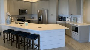 A modern kitchen with grey cabinets, a stainless steel refrigerator, a microwave, and a coffee maker. There are three bar stools at the island. - Enjoy Gorgeous Panoramic Views Of The Gulf Of Mexico And Vanderbilt Bay  (Naples)