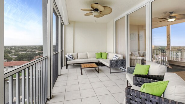 A balcony with a sofa, a coffee table, and a chair. There is a ceiling fan and a view of the city. - Enjoy Gorgeous Panoramic Views Of The Gulf Of Mexico And Vanderbilt Bay  (Naples)