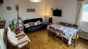 Een gezellige zithoek met een bank, eettafel en stoelen. Een televisie is aan de muur bevestigd, en er hangt een klok aan de muur.