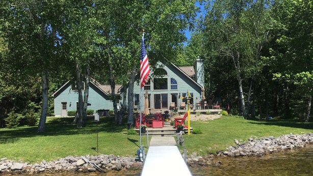 A house with a dock extending into a clear stream, surrounded by trees. -  Cozy, waterfront getaway for 2, w/loungers for relaxing & enjoying the sunsets (Lake Leelanau)