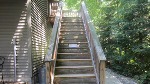A wooden staircase leading to a deck with a metal railing, surrounded by trees and gravel. -  Cozy, waterfront getaway for 2, w/loungers for relaxing & enjoying the sunsets (Lake Leelanau)