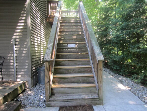 A wooden staircase leading to a deck with a metal railing, surrounded by trees and gravel.