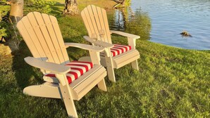 Two Adirondack chairs with red and white striped cushions, positioned on a grassy lakeside area with a view of the water and trees in the background. -  Cozy, waterfront getaway for 2, w/loungers for relaxing & enjoying the sunsets (Lake Leelanau)