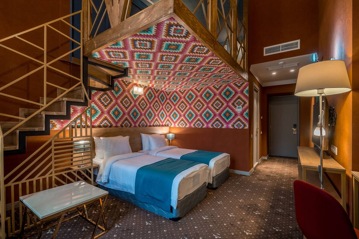 Habitación de hotel con cama, dos mesitas de noche, una silla, una escalera y una pared con un diseño geométrico.