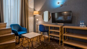 A hotel room with a TV, two blue chairs, a wooden desk, a lamp, and a mirror. - Hotel Gladius inn (Tbilisi)