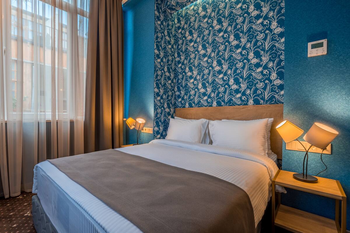 Una habitación de hotel con una cama, dos lámparas de noche, una mesita de noche y un ventanal con cortinas.