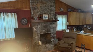 A cozy living room with a stone fireplace, a sofa, a coffee table with a lamp, and a kitchen area in the background. -  Pet friendly , Motorcycle parking  Hot tub. Close to everything (Maggie Valley)