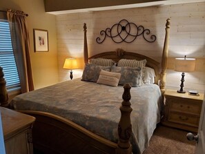 Un dormitorio con cama con dosel, cabecera de madera, un adorno en la pared, dos mesitas de noche con lámparas y un cajón junto a la cama.