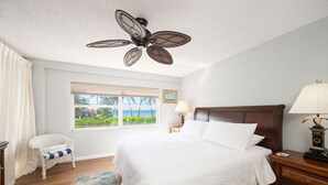 A hotel room with a large bed, a ceiling fan, a window with a view of the beach, a chair, and a nightstand with a lamp.