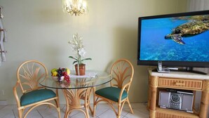 A dining area with a glass table, two wicker chairs, a potted plant, and a television displaying a sea turtle. - OceanFRONT, UPDATED, 4th Floor, 1BR Cali King, Non-Smoking, SHARP! (Kihei)