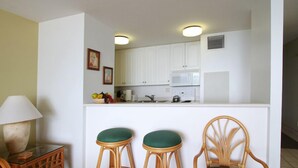 A compact kitchen with white cabinets, a microwave, and a small island with two stools. - OceanFRONT, UPDATED, 4th Floor, 1BR Cali King, Non-Smoking, SHARP! (Kihei)