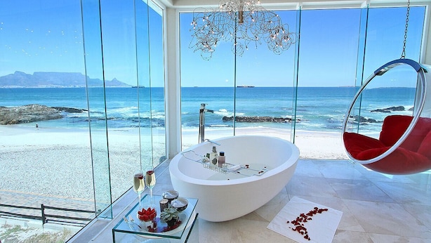 Une salle de bain moderne avec une grande baignoire, une table en verre où sont posés les articles de toilette, et un fauteuil suspendu près d’une grande fenêtre donnant sur une plage.