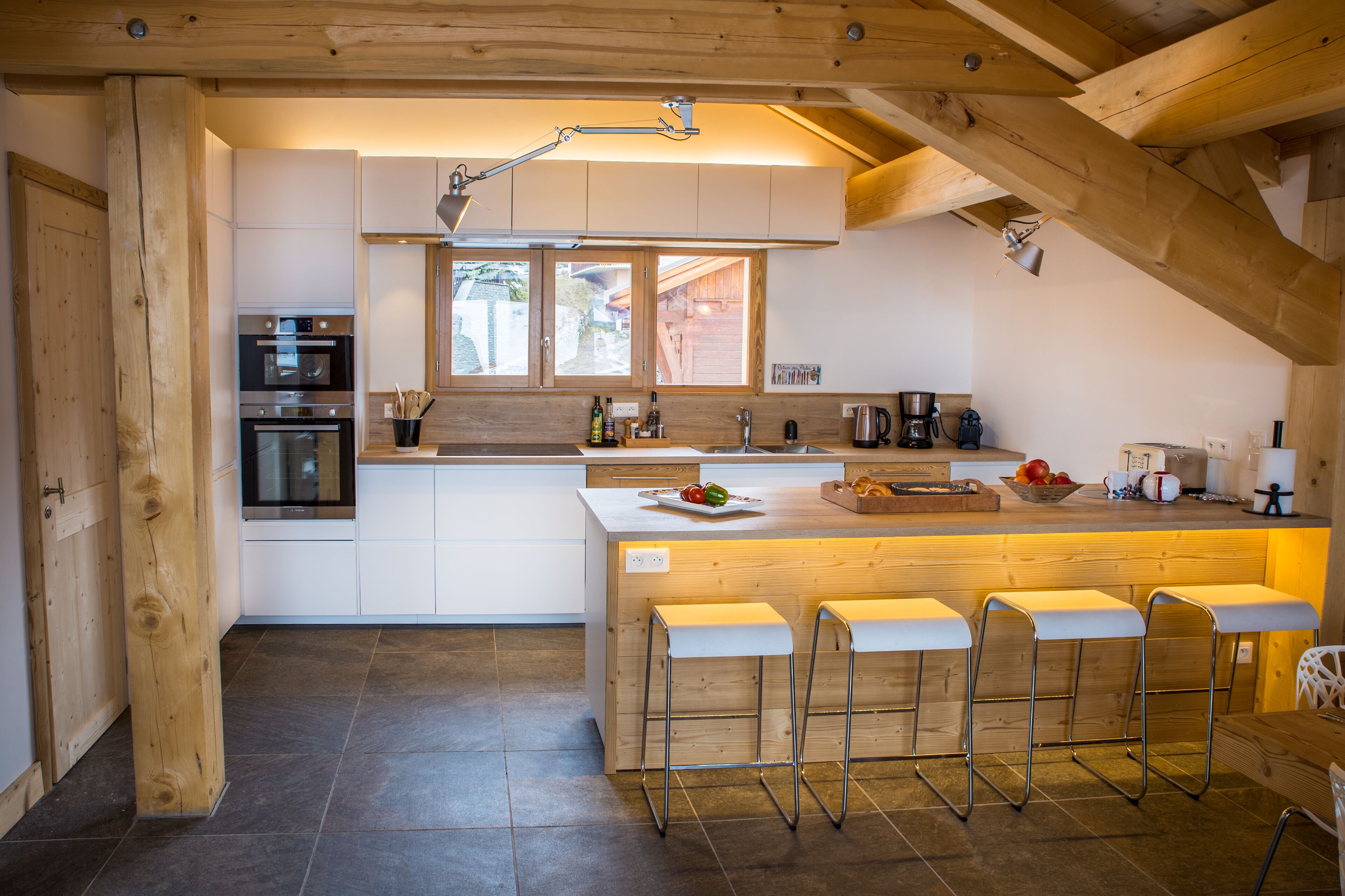Une cuisine moderne avec des poutres en bois, un îlot central doté de tabourets, des appareils électroménagers intégrés et une grande fenêtre.