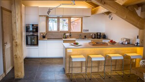 Een moderne keuken met houten balken, een centraal kookeiland met krukken, inbouwapparatuur en een groot raam.